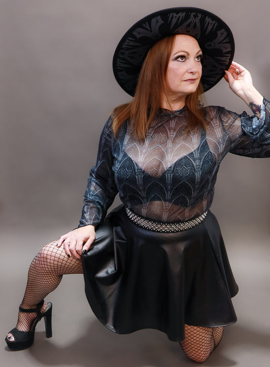 Woman wearing a black hat, sheer top, and black skirt with fishnet stockings against a gray background