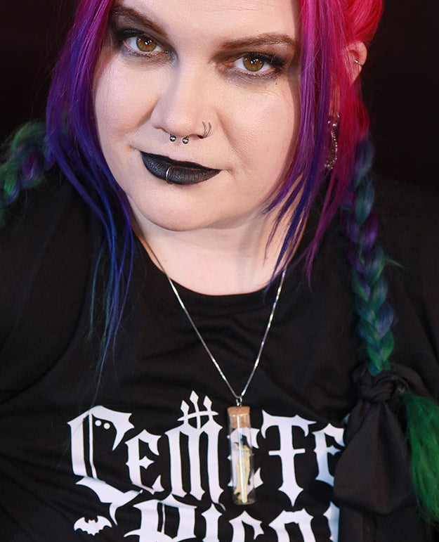 A person with bright pink, purple, and green hair poses in a black graphic t-shirt with 'Cemetery Picnic Club' printed in white gothic font, paired with fishnet stockings and a brown leather bracelet against a dark background.