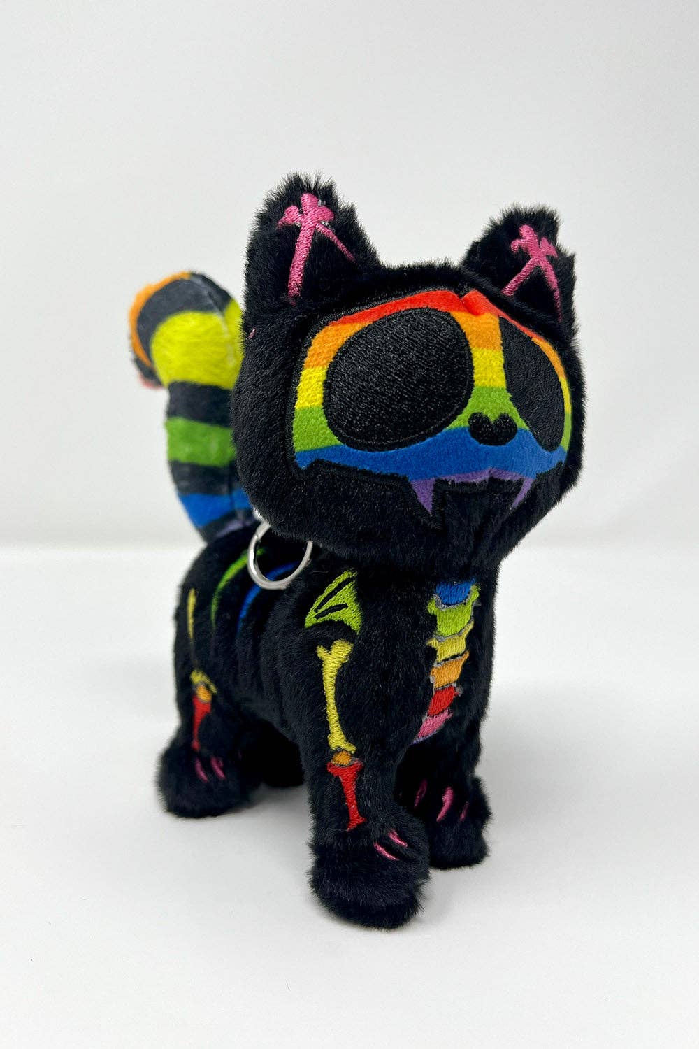 A black plush cat keychain with colorful embroidered details, including rainbow patterns around the eyes, pink X-shaped ears, and multicolor stripes on its tail and body, shown on a white background.