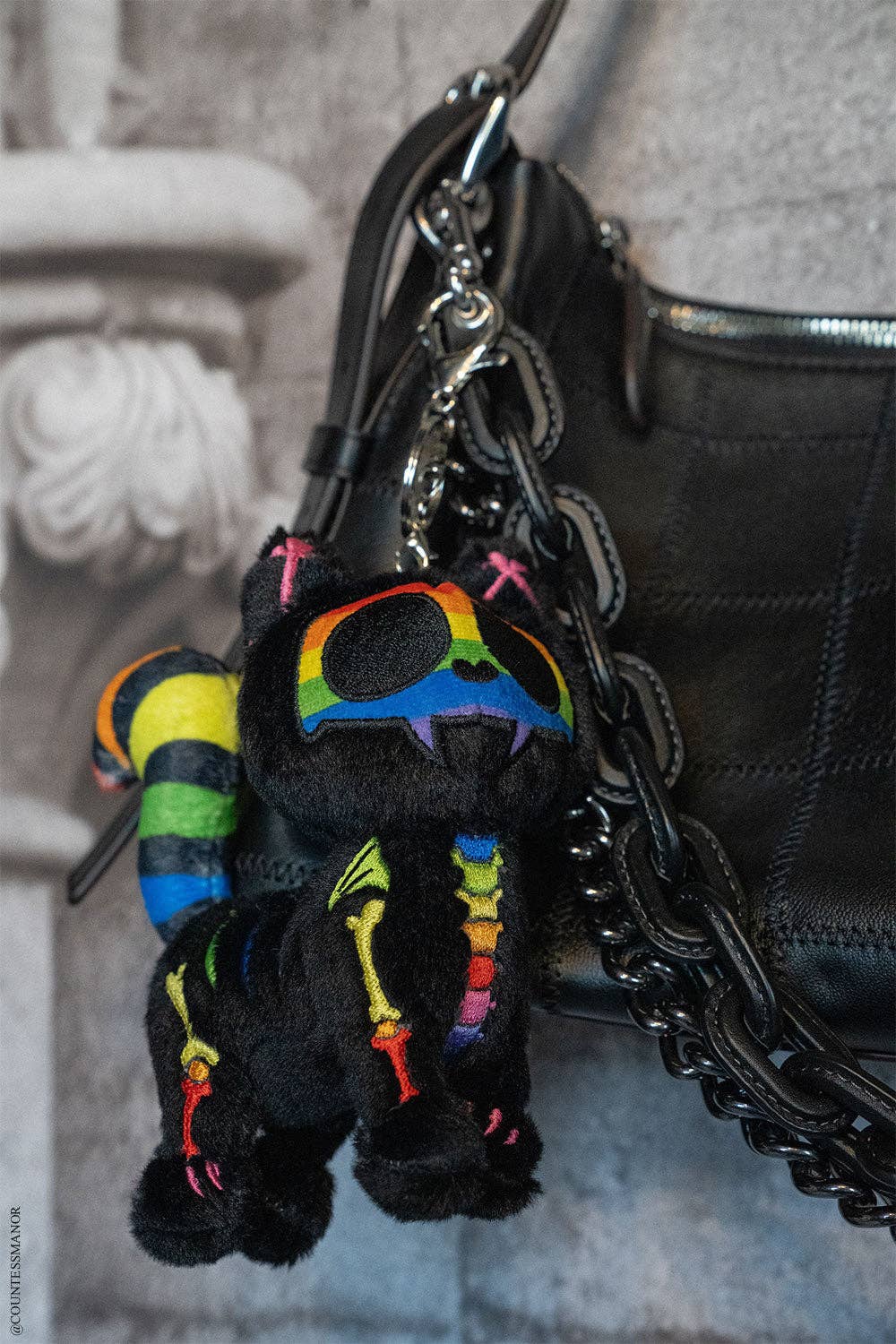 Black plush toy with rainbow accents attached to a black chain, hanging from a black leather bag.
