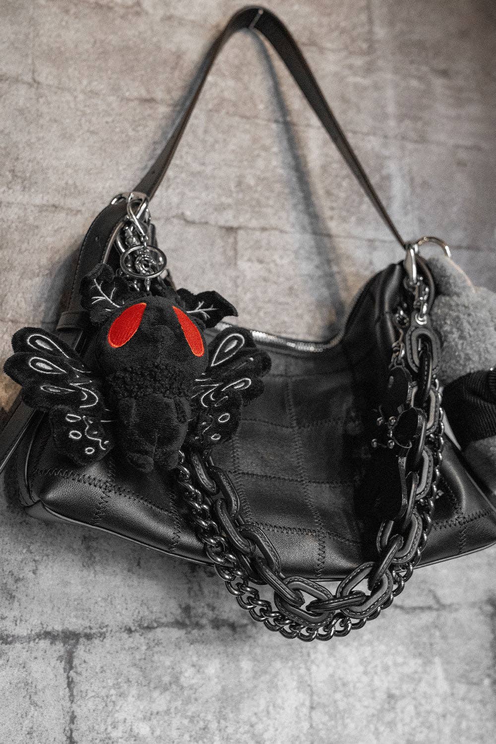 Black leather handbag with chain detail and a small moth man keychain plush on a textured surface.