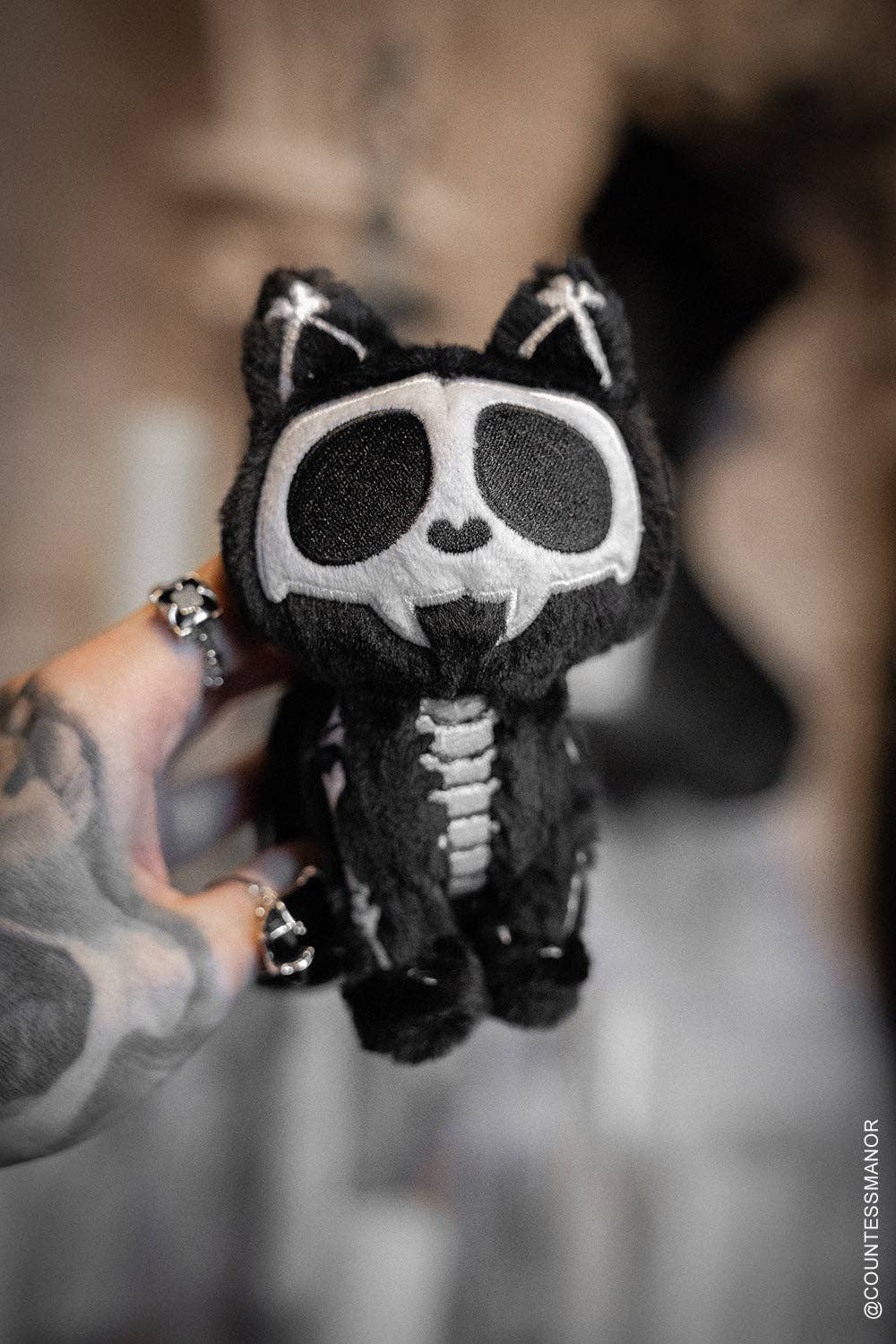 Black skeleton plush toy held by a person with tattoos against a blurred background