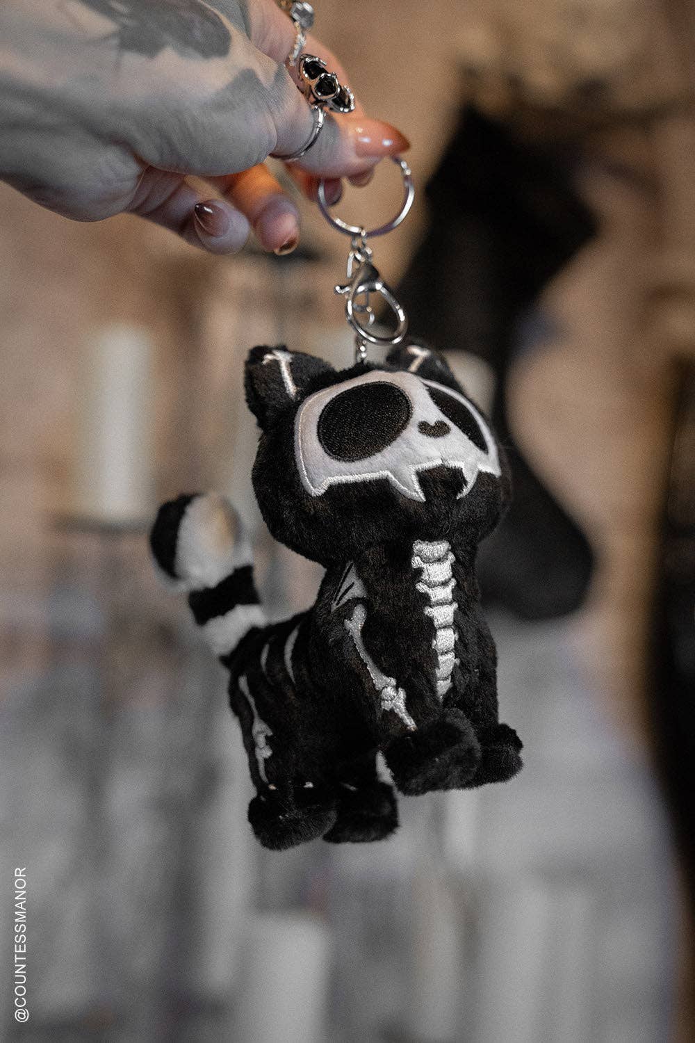 small plush cat keychain with a black-and-white skeleton design and silver key ring hardware being held against a blurred indoor background.
