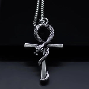 A silver-toned metal pendant necklace featuring an ankh symbol with a coiled snake wrapping through it, suspended from a beaded chain against a dark background.