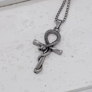 A silver-toned metal necklace featuring an ankh pendant with a coiled snake wrapped through it, resting on a textured white surface.