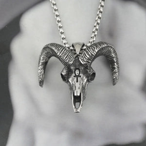 A silver-toned metal ram skull pendant necklace with curved horns and a ball chain, shown close up against a soft gray blurred background.
