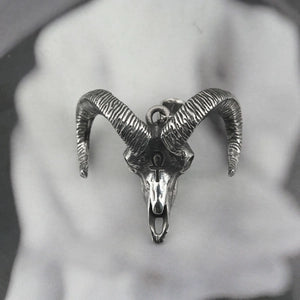 A detailed sterling silver ram skull pendant with curved horns, shown against a blurred black and white background.