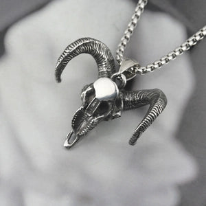 A silver-toned metal ram skull pendant necklace with curved horns, hanging from a matching chain against a soft gray blurred background.
