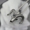 A detailed silver ram skull pendant necklace with curved horns, hanging from a silver chain against a soft, blurred gray background.