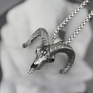 A detailed silver ram skull pendant necklace with curved horns, hanging from a silver chain against a soft, blurred gray background.
