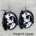 A pair of black and white oval dangle earrings featuring a gothic illustration of a kissing couple, shown against a textured gray background with text overlays indicating dimensions and weight.
