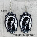 A pair of gothic-style oval dangle earrings with black and white illustrated portraits of a woman, shown against a light gray textured background. White text overlays indicate the size as 6.
