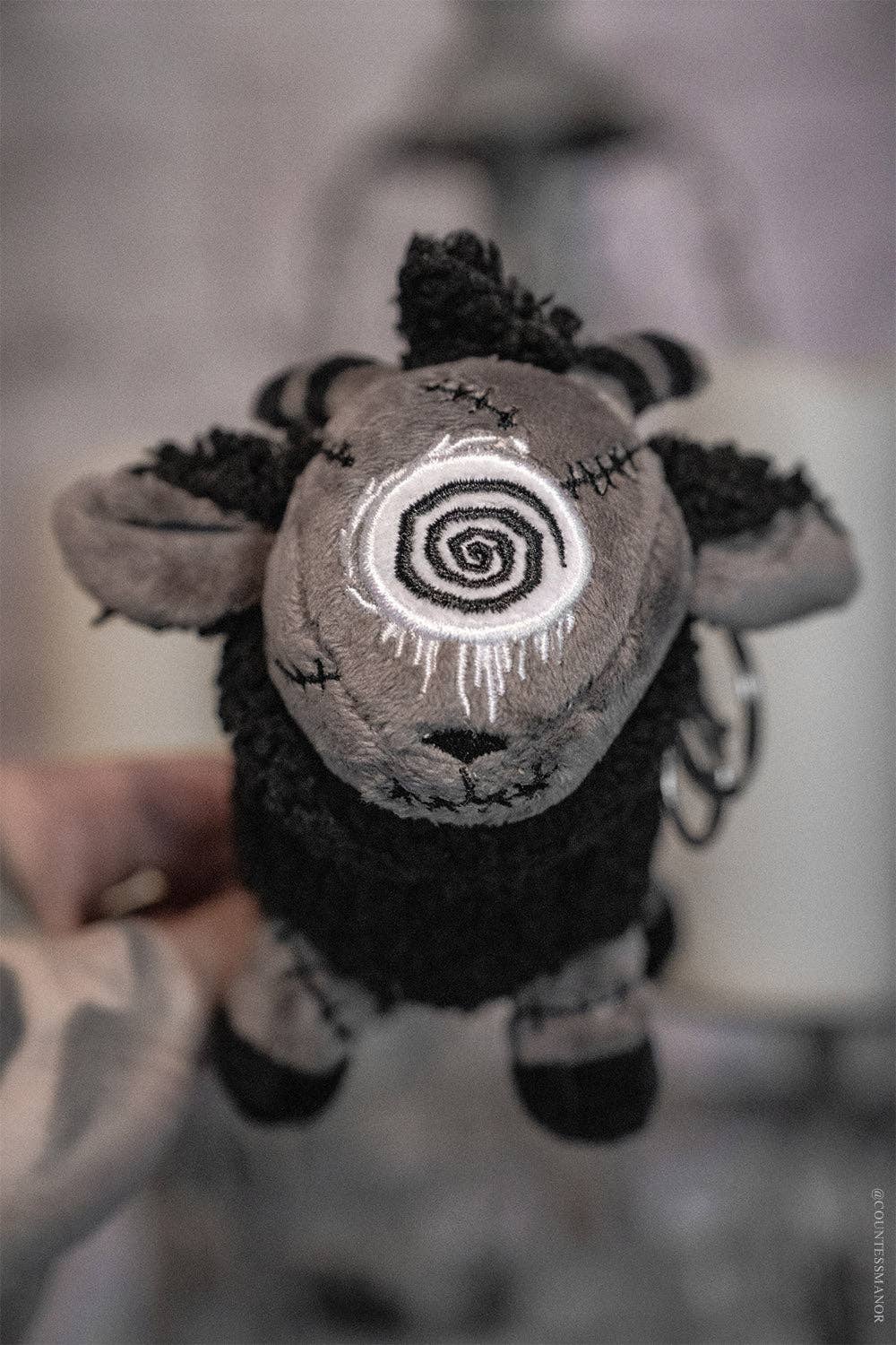 Black sheep plush keychain with a spiral design on its face held by a person against a blurred background