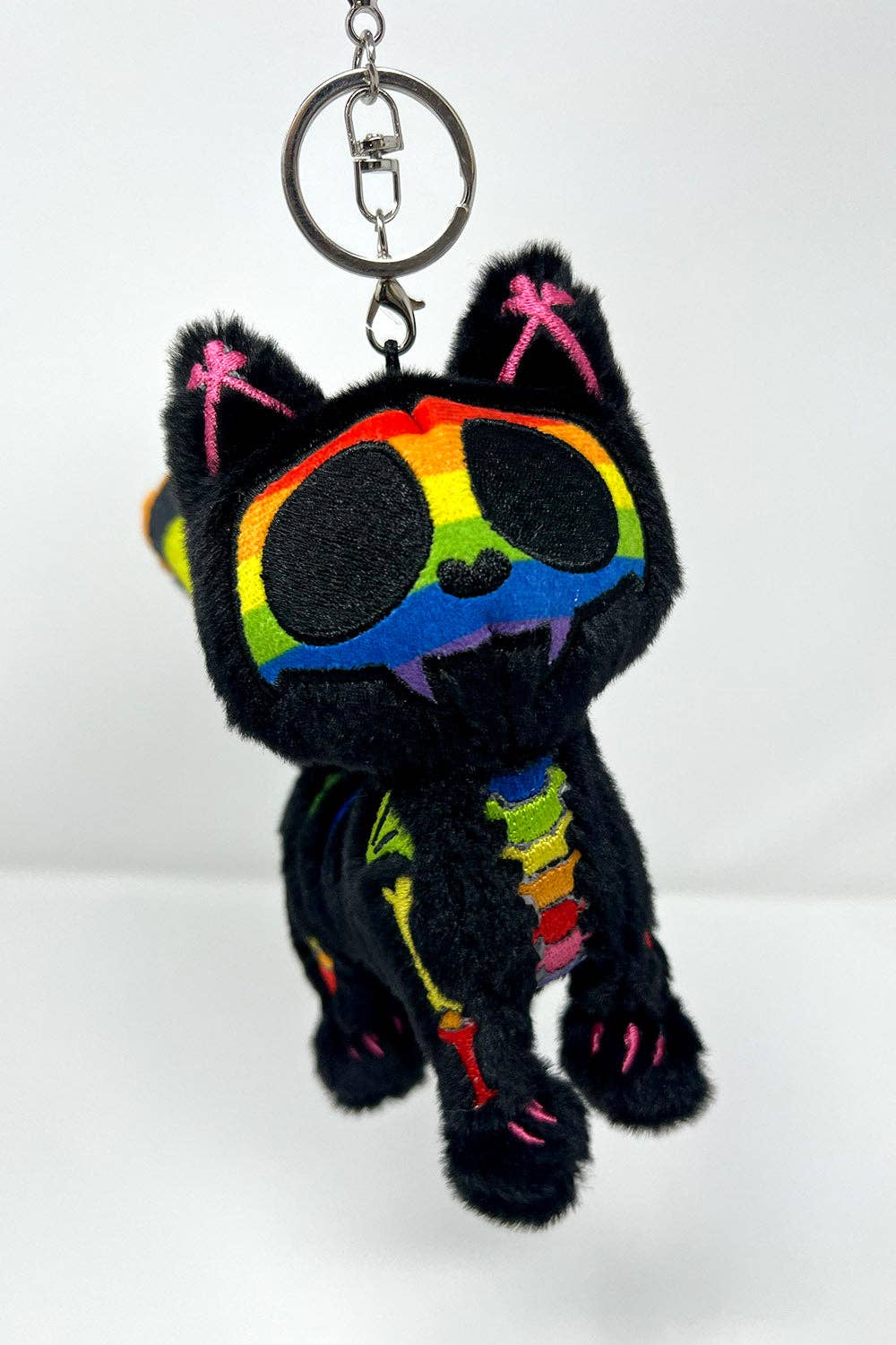 A black plush cat keychain with rainbow-colored embroidered details, including a rainbow around the eyes, pink accents on the ears and paws, and a rainbow-striped collar, shown against a plain white background.