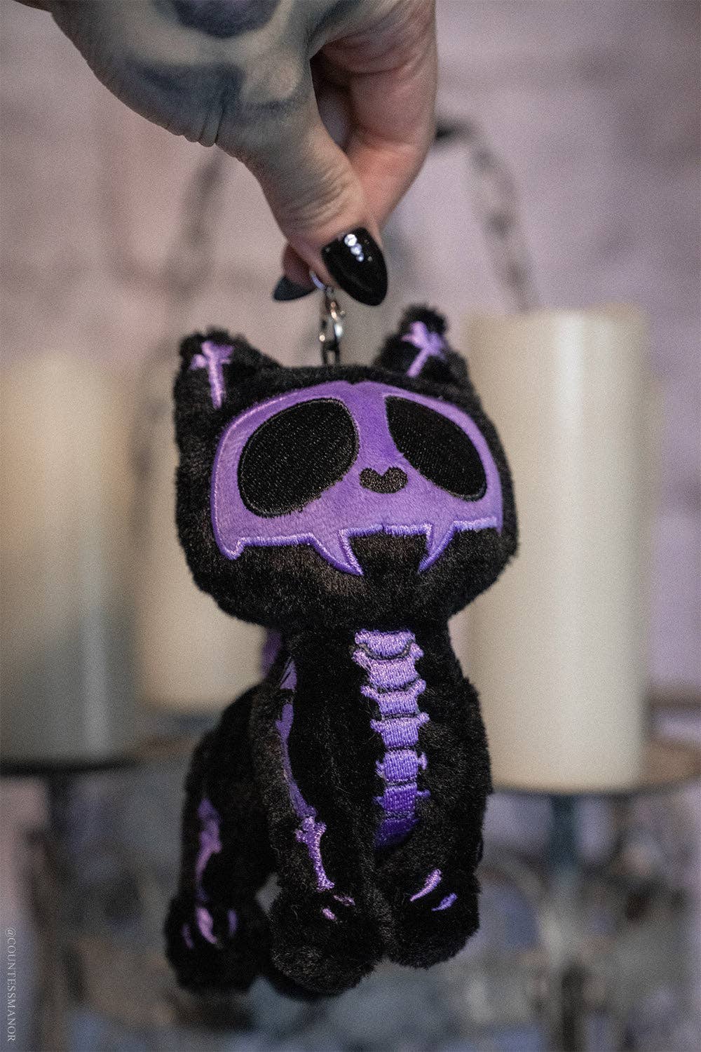 Black plush skeleton cat keychain with purple accents held by a hand against a blurred candlelit background.